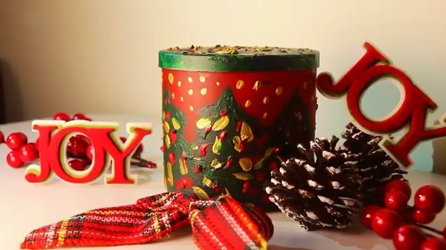 A red candle jar hand-painted with a Christmas tree, surrounded by festive decorations.